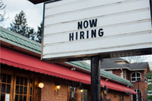 Typical american now hiring sign in black letters over the white background with local small business building 